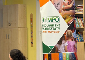 „Prowadząca warsztaty ekologiczne rozmawia z grupą dzieci siedzących na podłodze. Obok widoczne pojemniki do segregacji odpadów oraz roll-up z napisem: MPO Ekologiczne Warsztaty ‘Mini Wysypisko’.“