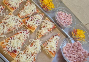 Bezglutenowa pizza przygotowana w kilku wariantach dietetycznych. Na blasze kawałki pizzy z sosem pomidorowym i startym serem, obok miseczki z dodatkami do samodzielnego komponowania porcji przez dzieci.