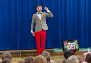 Mężczyzna w szarej marynarce, zielonej muszce i czerwonych spodniach stoi na scenie przed niebieską kurtyną i występuje jako iluzjonista dla grupy dzieci siedzących na widowni. W jednej ręce trzyma czerwoną piłeczkę, a drugą unosi w górę w dynamicznym geście. Obok niego stoi czarna walizka z napisem „Magik Konrad.pl”.