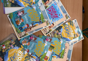 Na drewnianym stole rozłożone są książki z serii „Dobre bajki” autorstwa Małgorzaty Korbiel oraz dyplomy. Kolorowe okładki przedstawiają ilustracje dzieci, zwierząt i przyrody. Książki ułożone są w kilku egzemplarzach, przygotowane jako nagrody dla uczestników konkursu.