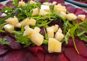 Na talerzach ułożone cienkie plastry buraka tworzące carpaccio, udekorowane świeżą rukolą oraz kostkami soczystej gruszki. W tle widoczne kolejne porcje dania przygotowane dla dzieci.