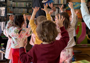 Grupa dzieci z przedszkola uczestniczy w zajęciach edukacyjnych w bibliotece. Dzieci stoją na kolorowych poduszkach i podnoszą ręce do góry podczas zabawy prowadzonej przez bibliotekarza, który trzyma książkę. W tle widać regały pełne książek i przyjazną przestrzeń biblioteczną. Zajęcia odbyły się w Bibliotece nr 10 przy ul. Anielewicza 2, z udziałem dzieci z grupy II „Wilczki”.