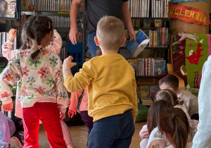 Grupa dzieci z przedszkola uczestniczy w zajęciach edukacyjnych w bibliotece. Dzieci stoją na kolorowych poduszkach i podnoszą ręce do góry podczas zabawy prowadzonej przez bibliotekarza, który trzyma książkę. W tle widać regały pełne książek i przyjazną przestrzeń biblioteczną. Zajęcia odbyły się w Bibliotece nr 10 przy ul. Anielewicza 2, z udziałem dzieci z grupy II „Wilczki”.
