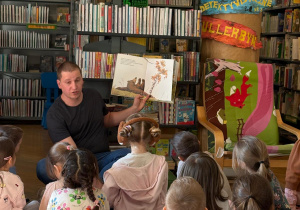 Grupa dzieci z przedszkola uczestniczy w zajęciach edukacyjnych w bibliotece. Dzieci stoją na kolorowych poduszkach i podnoszą ręce do góry podczas zabawy prowadzonej przez bibliotekarza, który trzyma książkę. W tle widać regały pełne książek i przyjazną przestrzeń biblioteczną. Zajęcia odbyły się w Bibliotece nr 10 przy ul. Anielewicza 2, z udziałem dzieci z grupy II „Wilczki”.