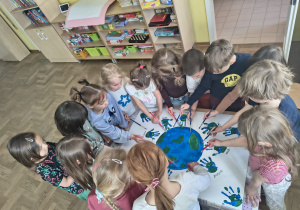 „Grupa dzieci pracuje razem nad plakatem przedstawiającym Ziemię, malując ją farbami i tworząc wokół niej odciski dłoni.”