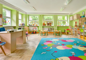 Kolorowa sala przedszkolna z żółtymi ścianami, stolikami i krzesełkami oraz dywanem z planetą.