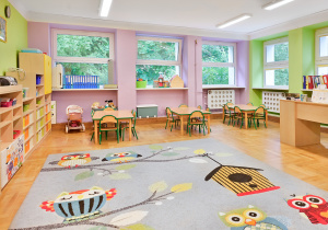 Kolorowa sala przedszkolna z żółtymi ścianami, stolikami i krzesełkami oraz dywanem z planetą.