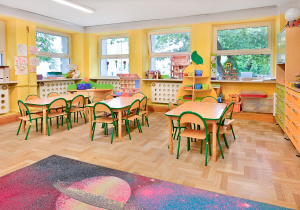 Kolorowa sala przedszkolna z żółtymi ścianami, stolikami i krzesełkami oraz dywanem z planetą.
